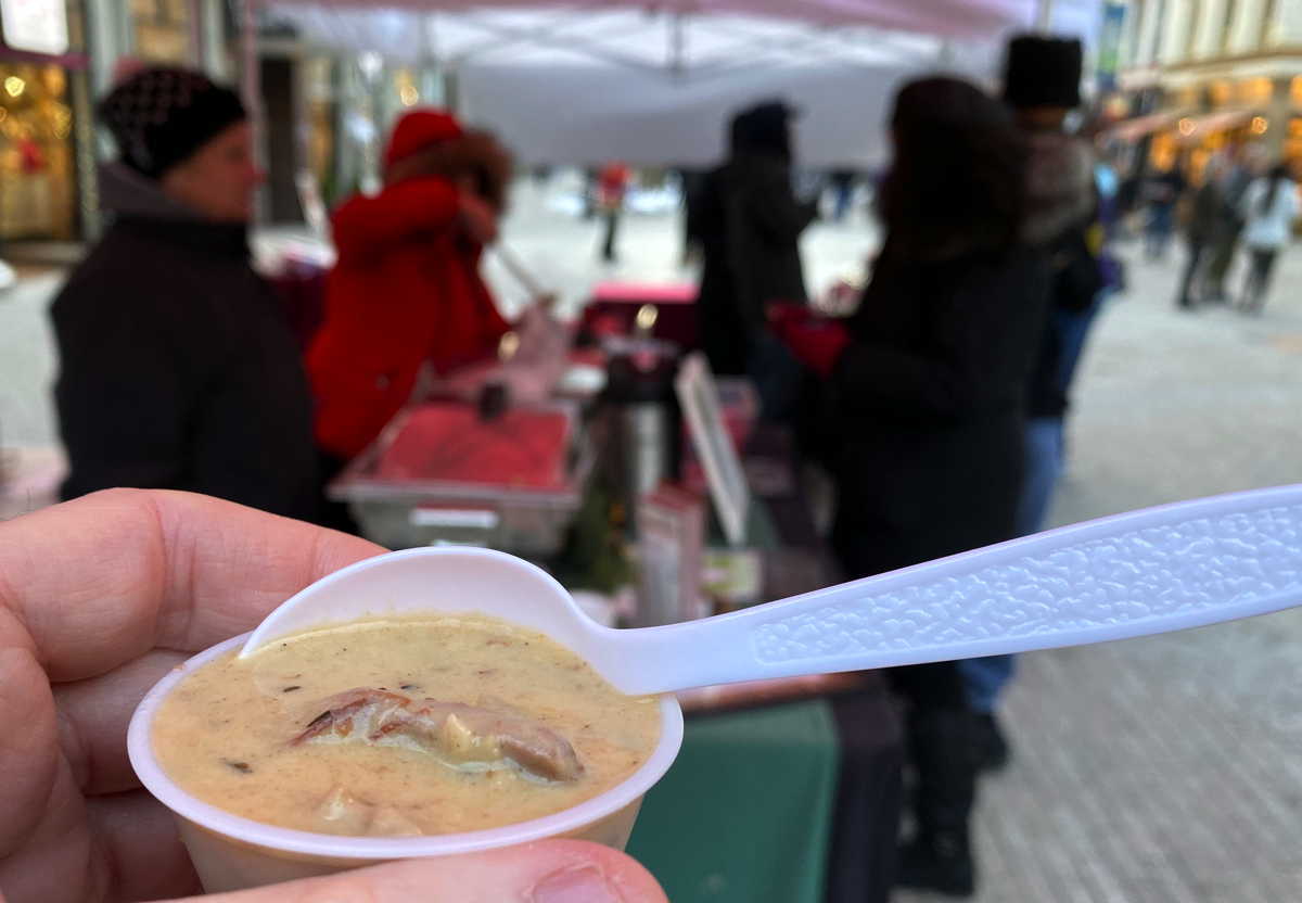 A tasting cup of chowder with a spoon sticking out, with people at an event booth in the background