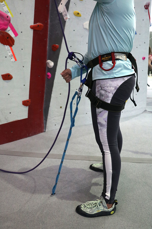 A belayer has clipped the ground anchor to their belay loop, below the separately clipped belay device