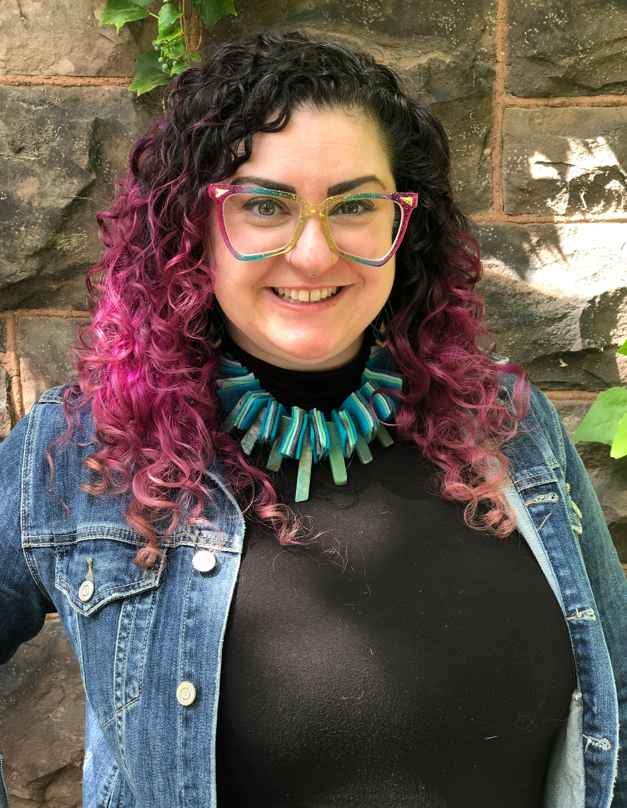 A smiling white woman with purple hair and rainbow glasses stands in front of a stone wall