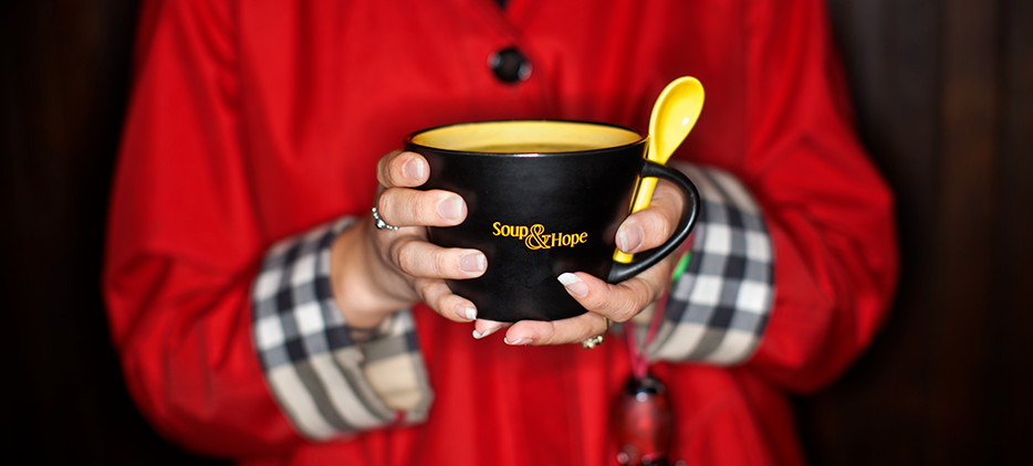 A person in a red coat holds a Soup & Hope bowl and spoon.