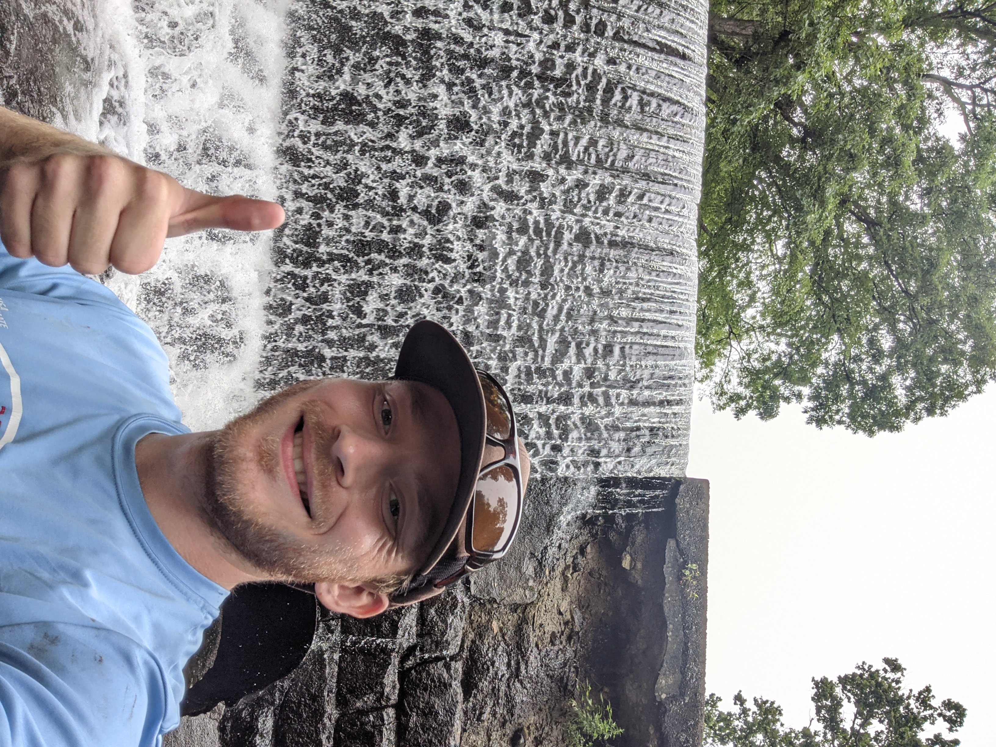 Student stands, giving a thumbs up, in front of a waterfall.