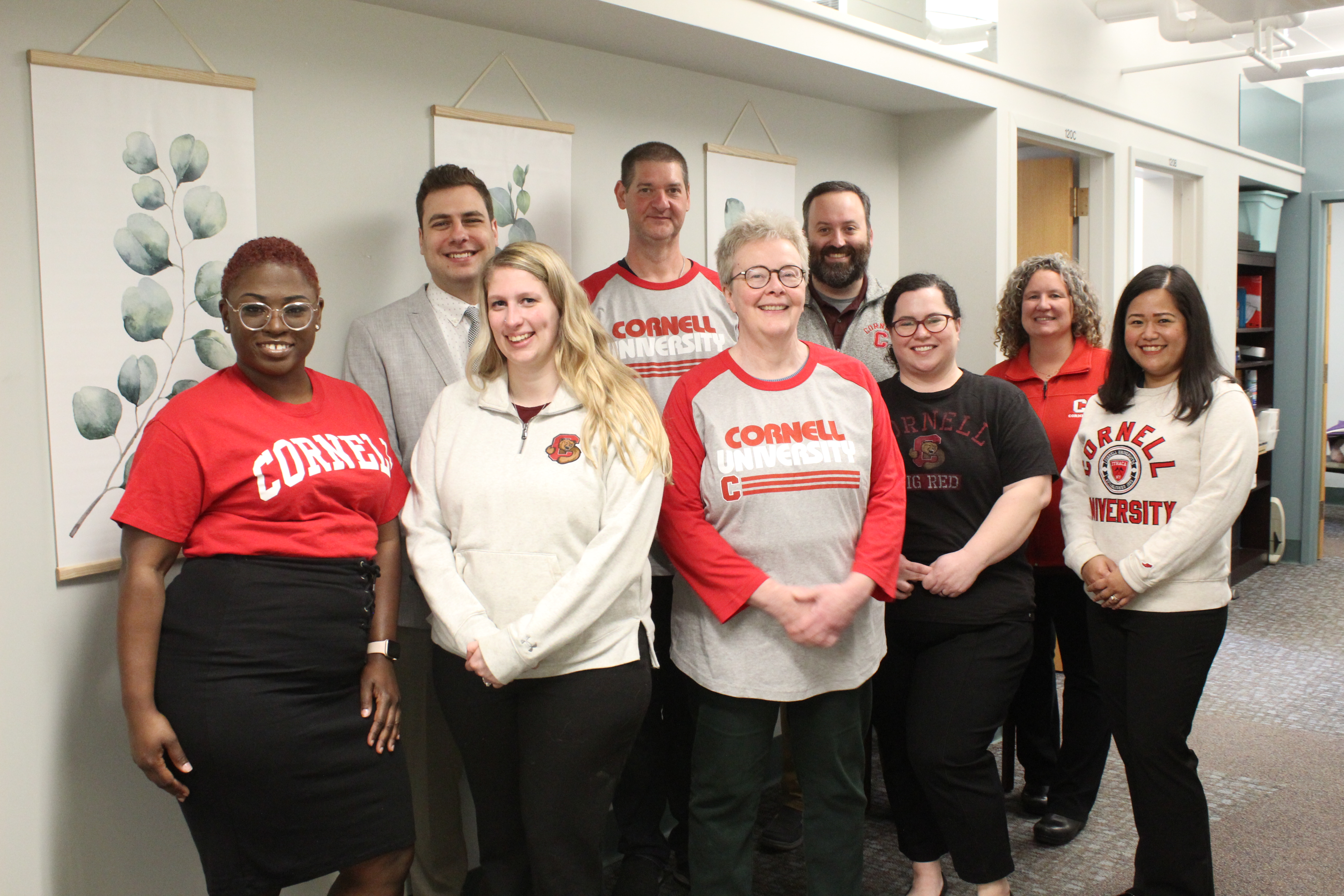 Nine members of the Office of Student Conduct and Community Standards pose for a group photo in front of a white wall wearing red Cornell gear.