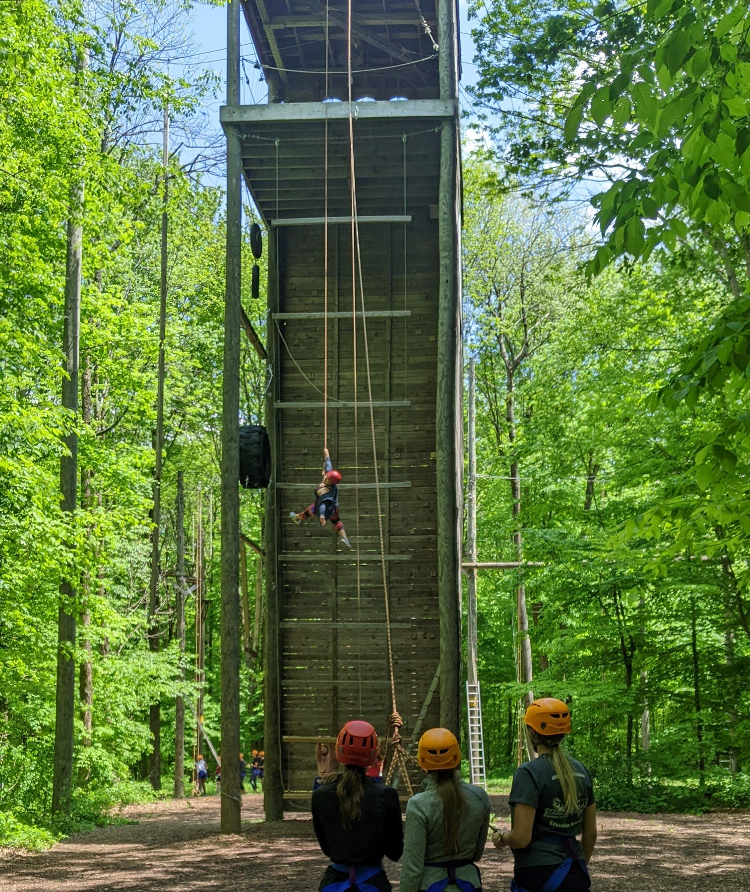 Participant descending from tower at ropes course, while being belayed by three people on the ground in the foreground of the image. 