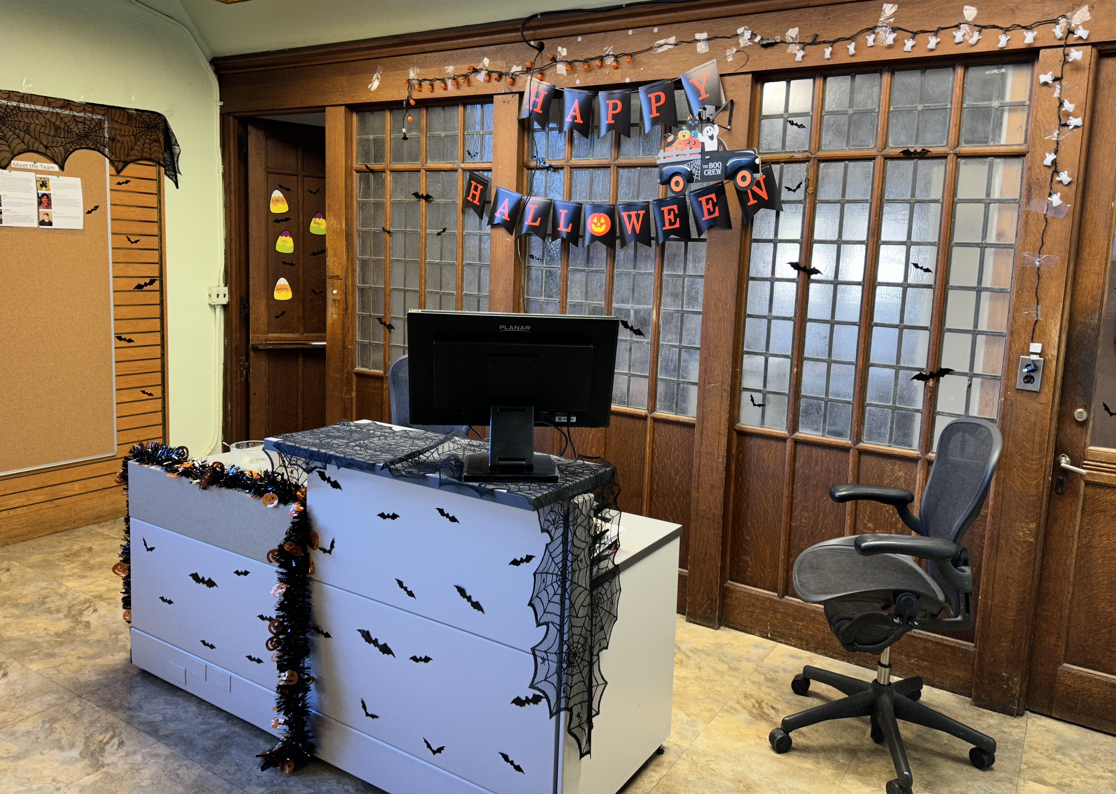 A desk and rolling office chair in front of a wood and frosted glass wall. Halloween decorations include a Happy Halloween banner on the wall, and a spiderweb drape and paper bats on the desk