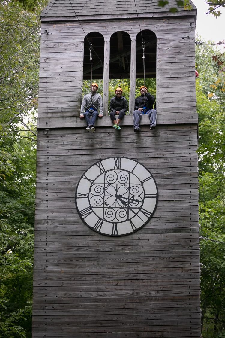 three people sit in the three windows of a wooden tower, attached to zip lines. There is a clock with roman numerals painted on the tower underneath them.