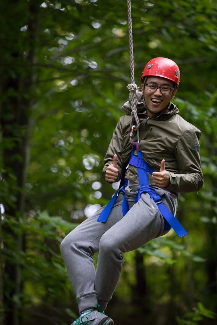 Man in red helmet and glasses with big smile gives double thumbs up from zip line