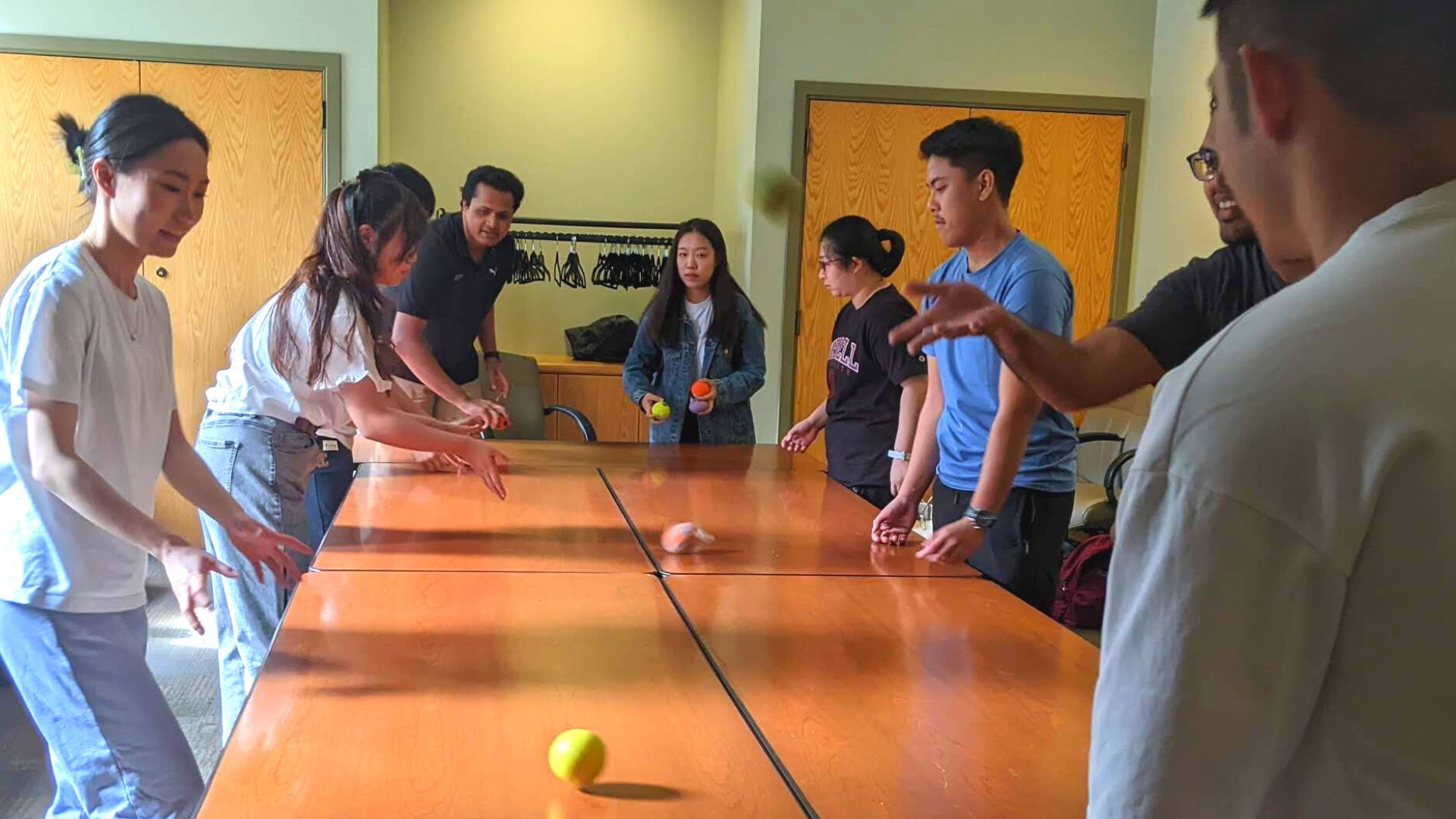 Students gathered around a wooden table passing tennis balls across the table while looking very focused and smiling.