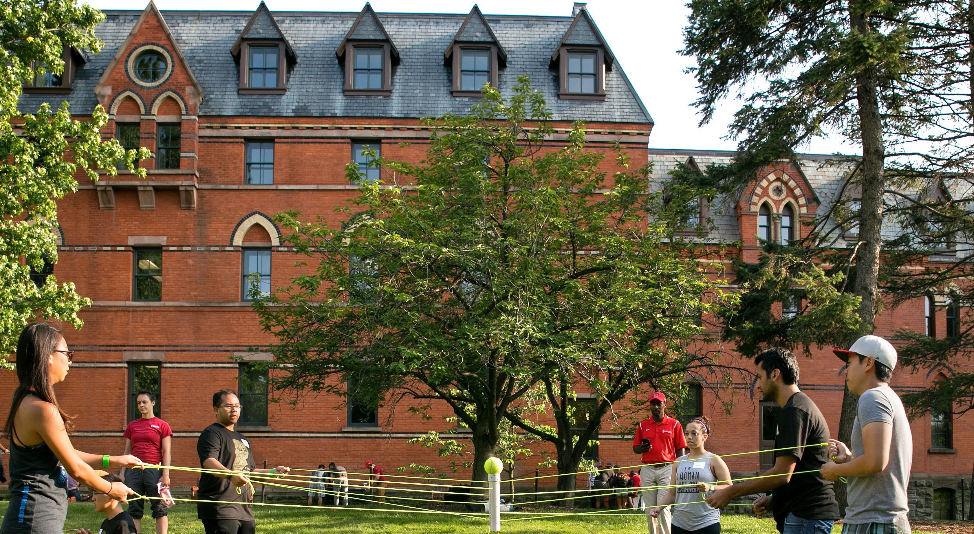 Students in front of brick building holding onto a string that connects to a balanced ball in the center 