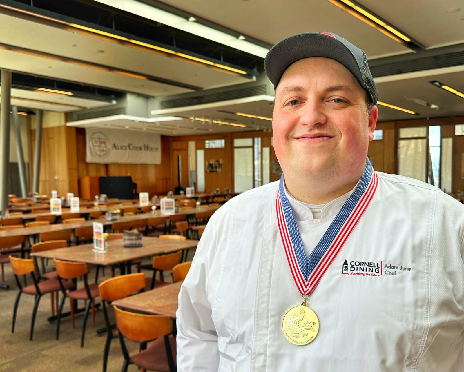 A smiling chef stands in front of a residential dining room wearing a chef coat, a cap, and a gold medal on a ribbon around his neck