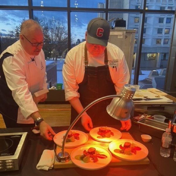 Two chefs arrange four plates of food under a warming lamp