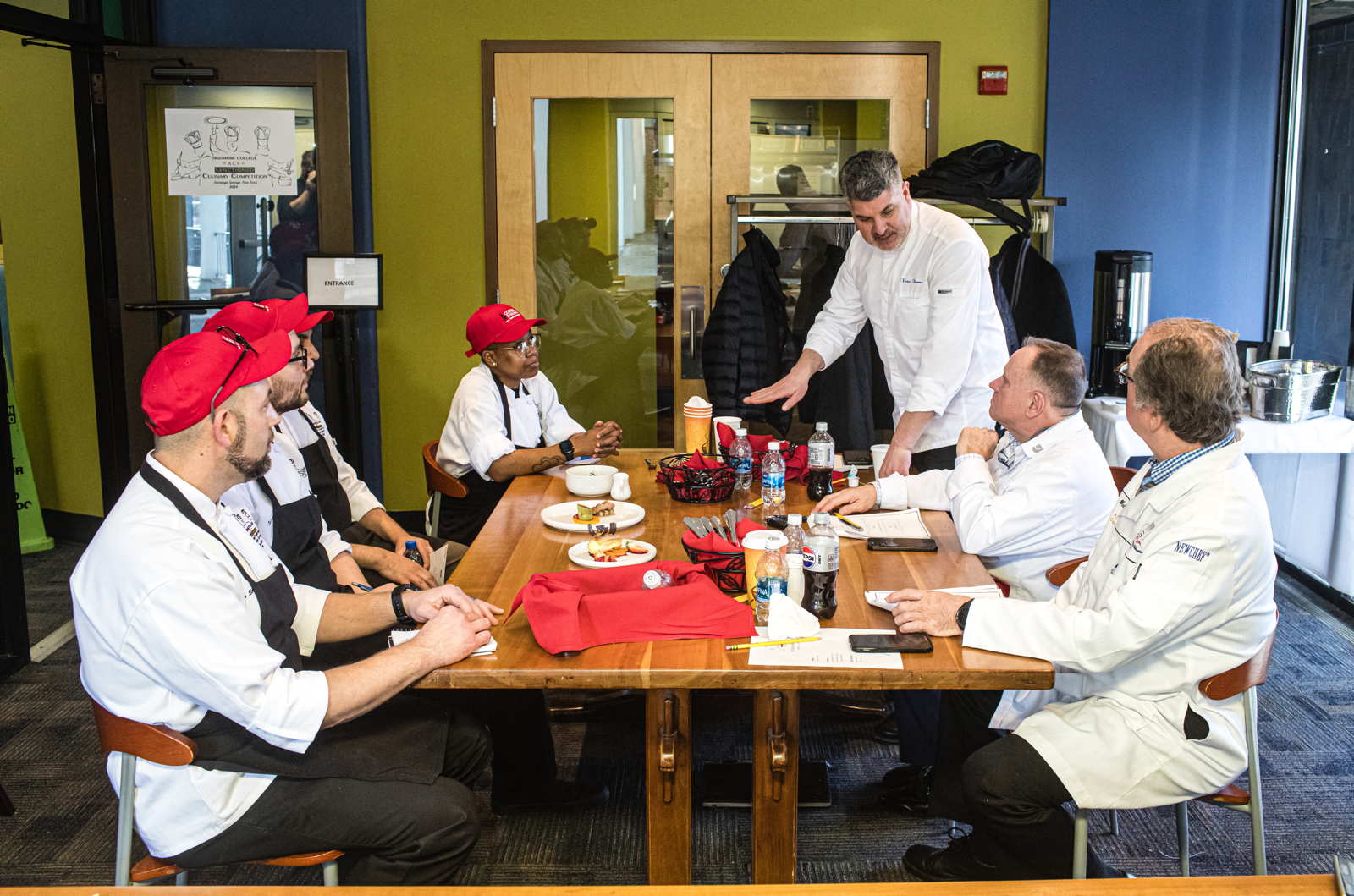 Several people wearing chef coats sit around a table while another stands at the corner of the table