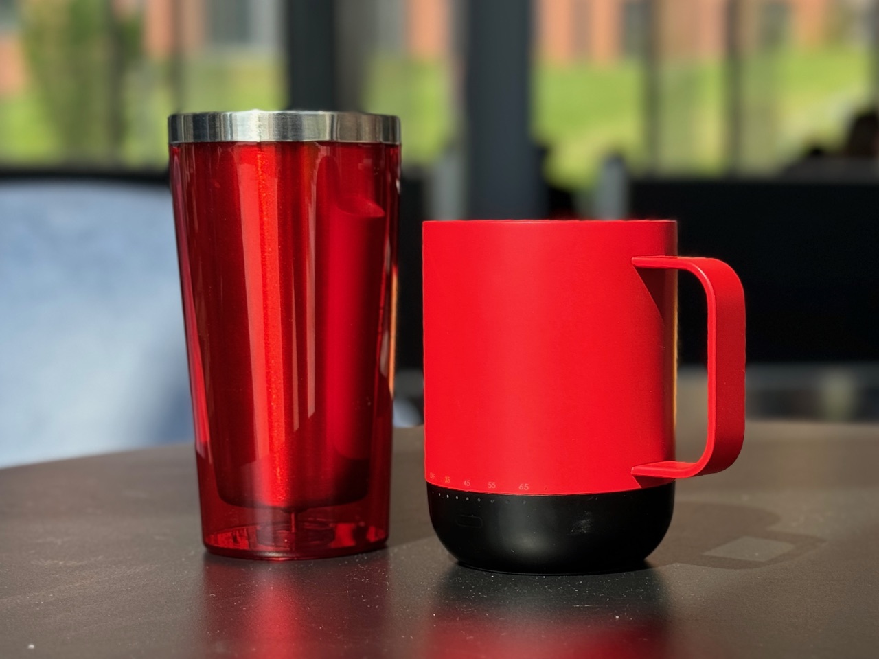 A red reusable tumbler and a red and black reusable mug sit on a cafe table.