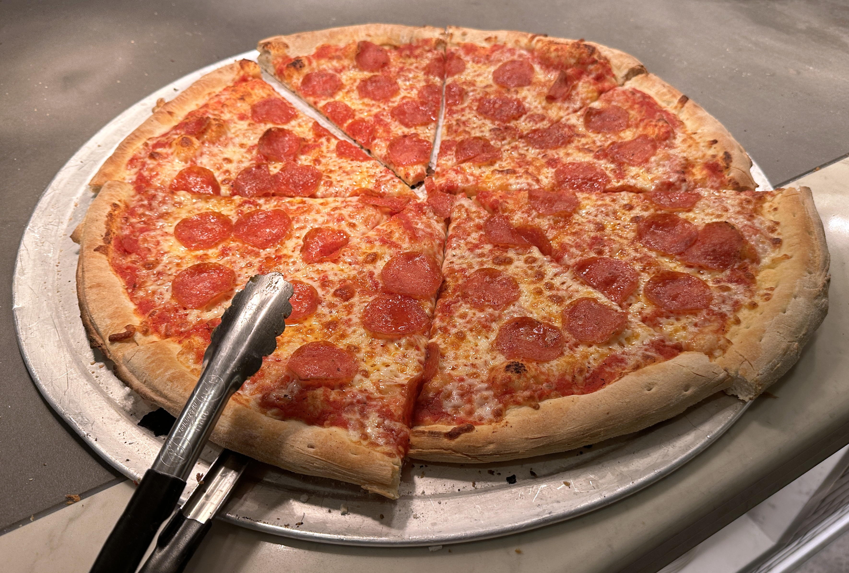 Tongs reach for a slice of pizza out of a pepperoni pizza resting on a pan