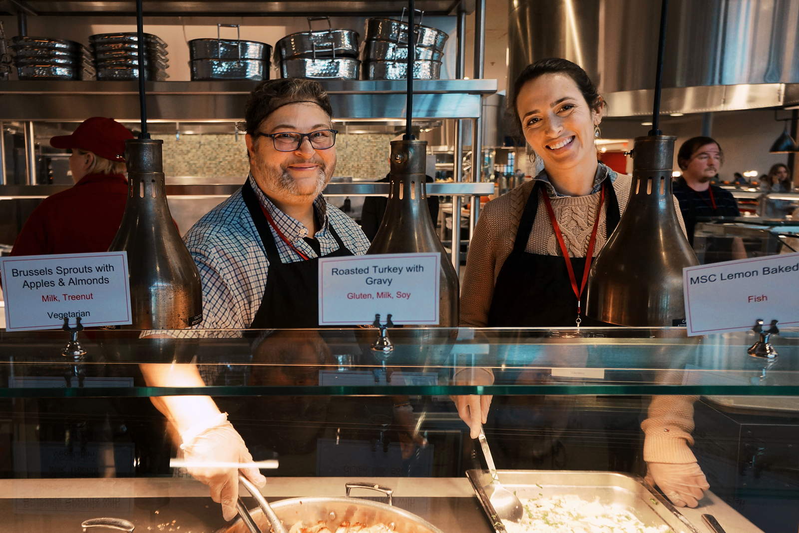 Two smiling people stand behind a food serving line