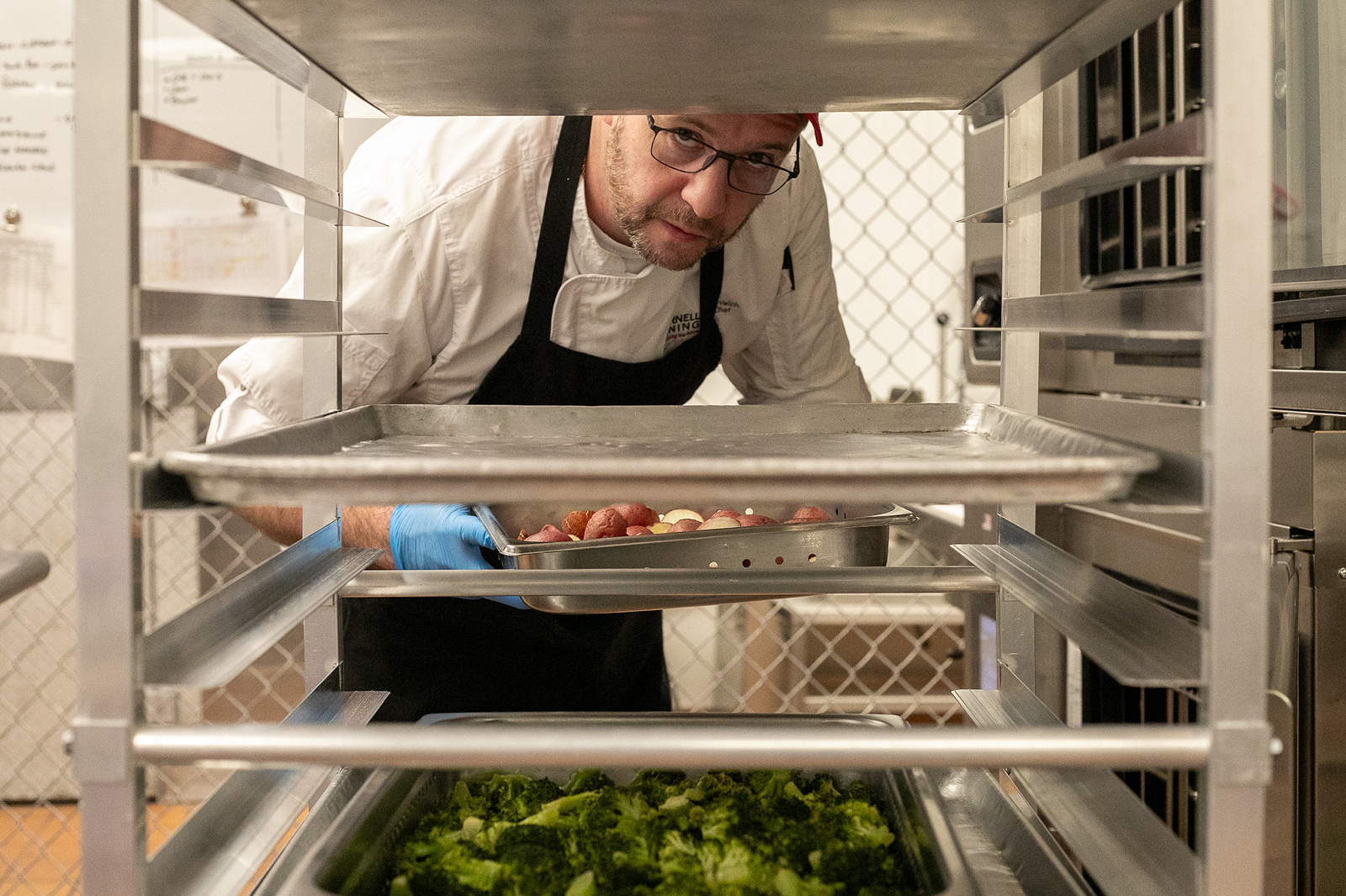 A person in a chef coat and apron peers through a rack holding trays of cooked food