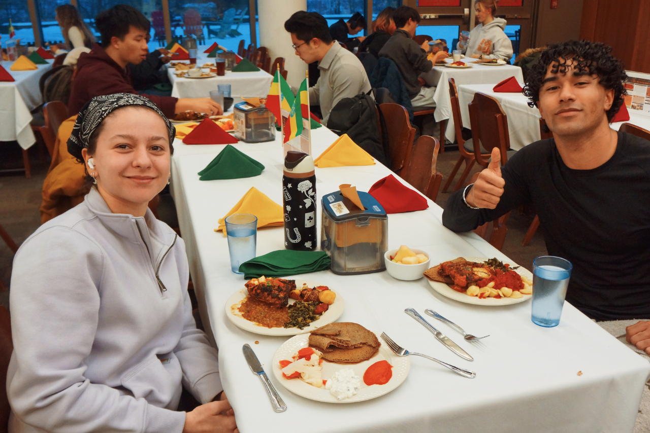 Students on either side of a dining room table with plates of food, one giving a thumbs up