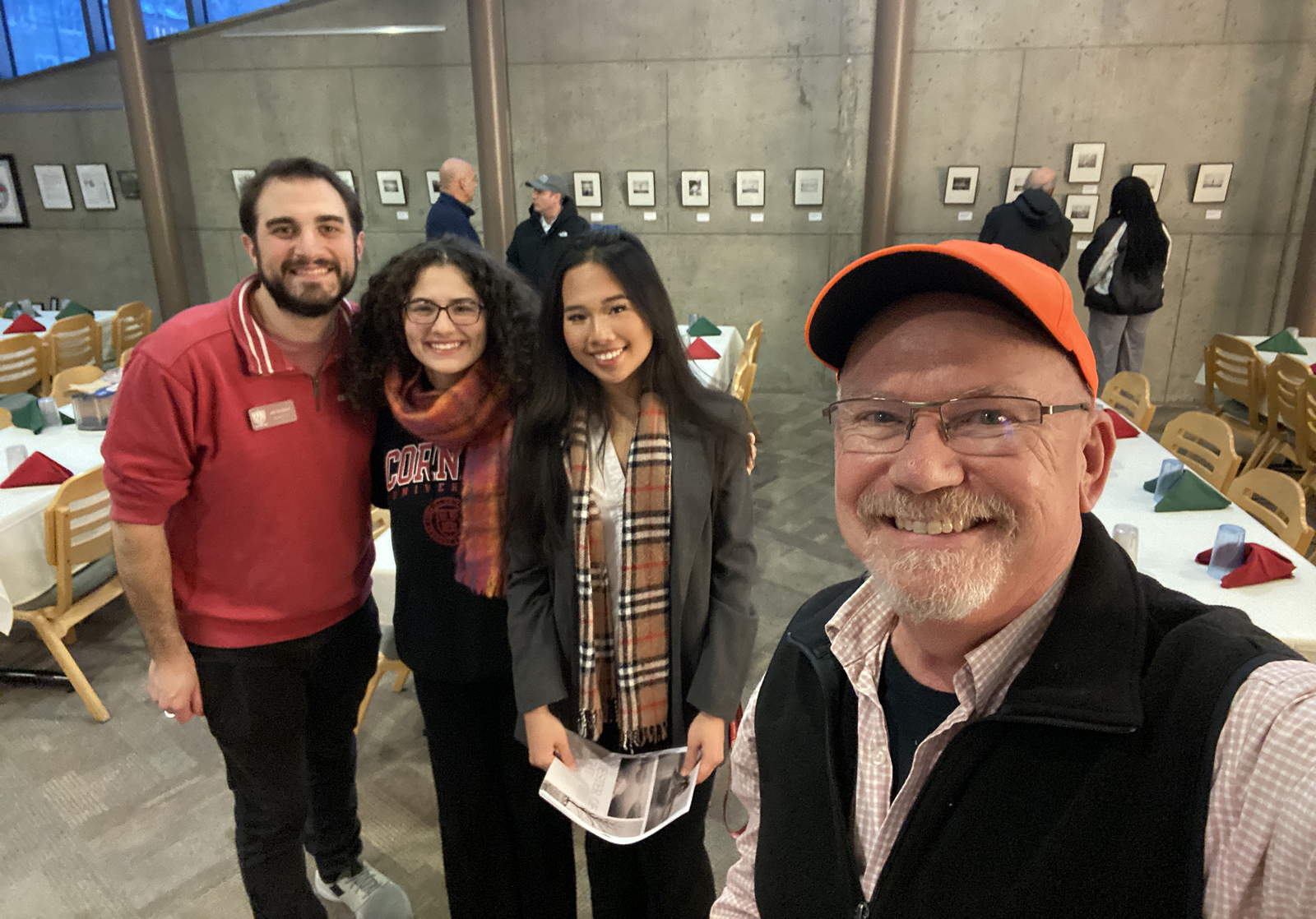 A smiling person in the foreground takes a selfie with a trio of people just behind him. In the background, more people stand in front of a row of photos on the wall.