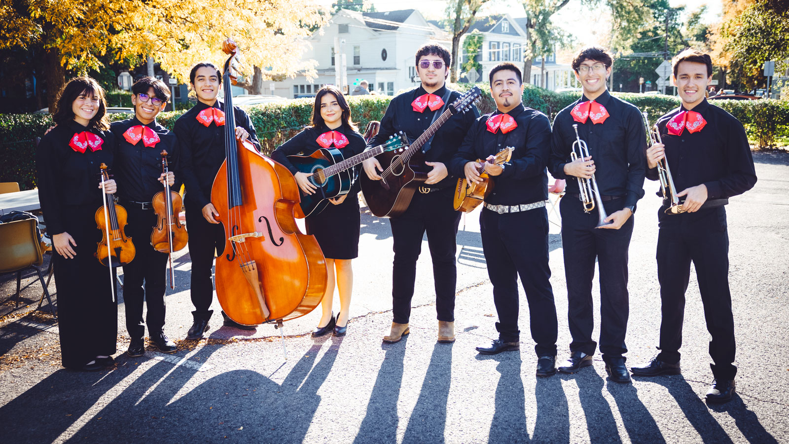 Eight-member mariachi band in traditional black uniforms with red bow ties poses outdoors, holding various instruments including strings and brass, with autumn trees and buildings behind them.