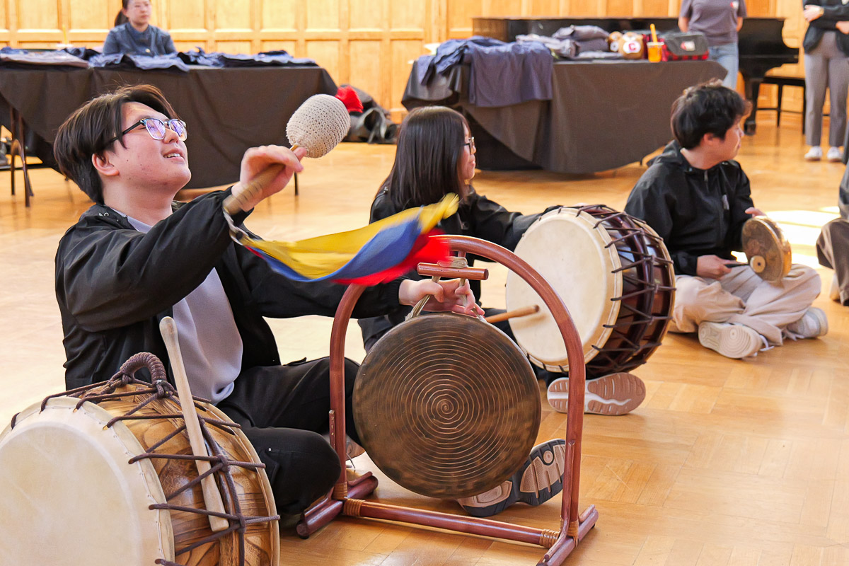 A student with glasses sits cross-legged on a wooden floor, playing a circular gong mounted on a stand while holding a beater. They wear dark clothing with a colorful ribbon on their arm. Other students with traditional percussion instruments, including drums, sit nearby in what appears to be a music workshop at Cornell University.