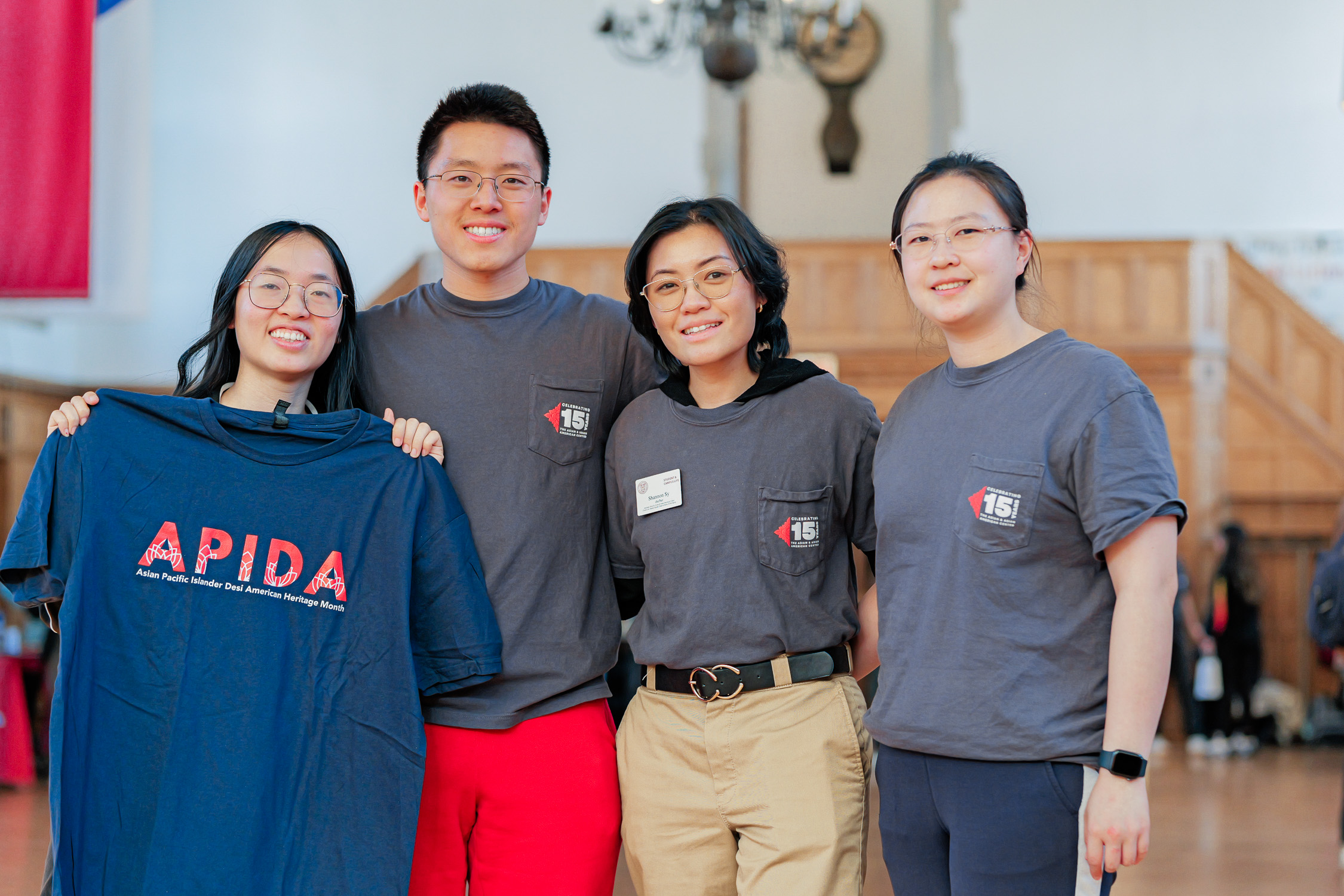 Four individuals pose together smiling at the A3C APIDA Heritage Month launch event, one holding a t-shirt that reads “APIDA: Asian Pacific Islander Desi American Heritage Month.” Others wear shirts commemorating A3C’s 15th anniversary.