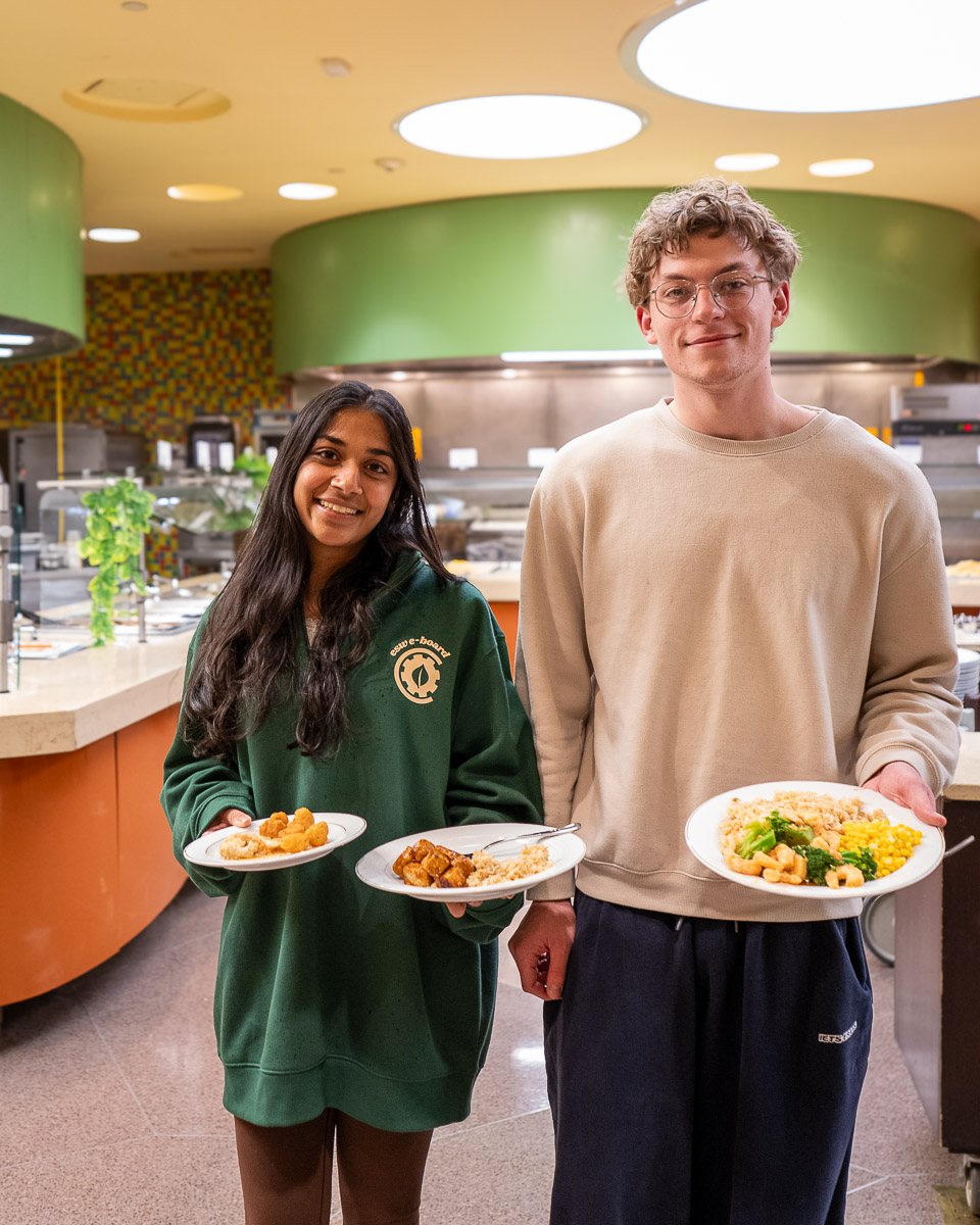 Two smiling students stand in a modern dining hall holding plates of food. A woman in a green Cornell sweatshirt holds a plate with what appears to be fried items, while a tall man with glasses and curly hair in a beige sweater holds a plate with rice, vegetables, and other dishes. The background shows a contemporary cafeteria with green walls, circular ceiling lights, and food service areas.