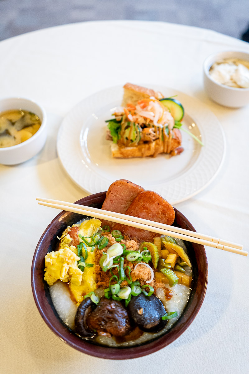 Bowl of congee topped with Spam, scrambled eggs, mushrooms, pickled vegetables, scallions, and pork floss, with miso soup, yogurt, and a bánh mì sandwich on a white table.