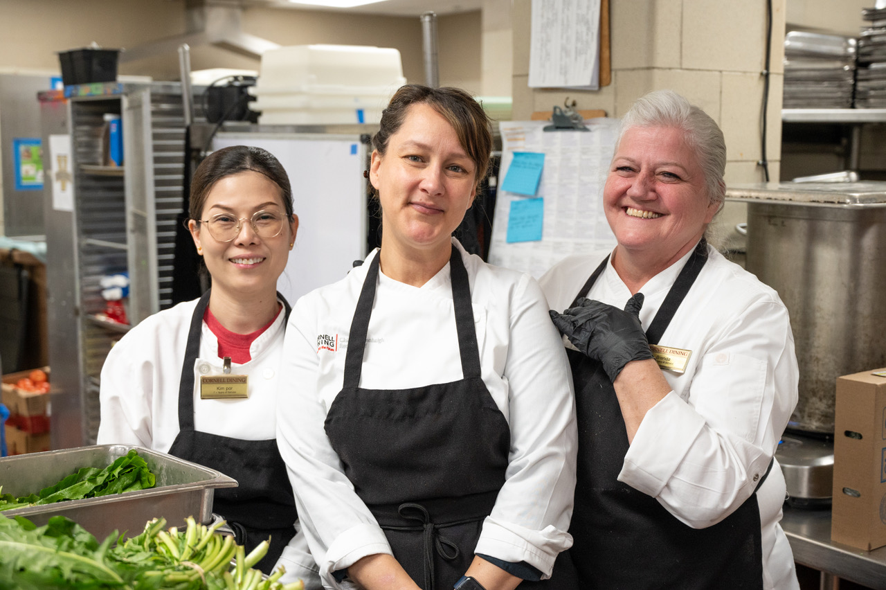 Three women in chef coats and black aprons in a college kitchen