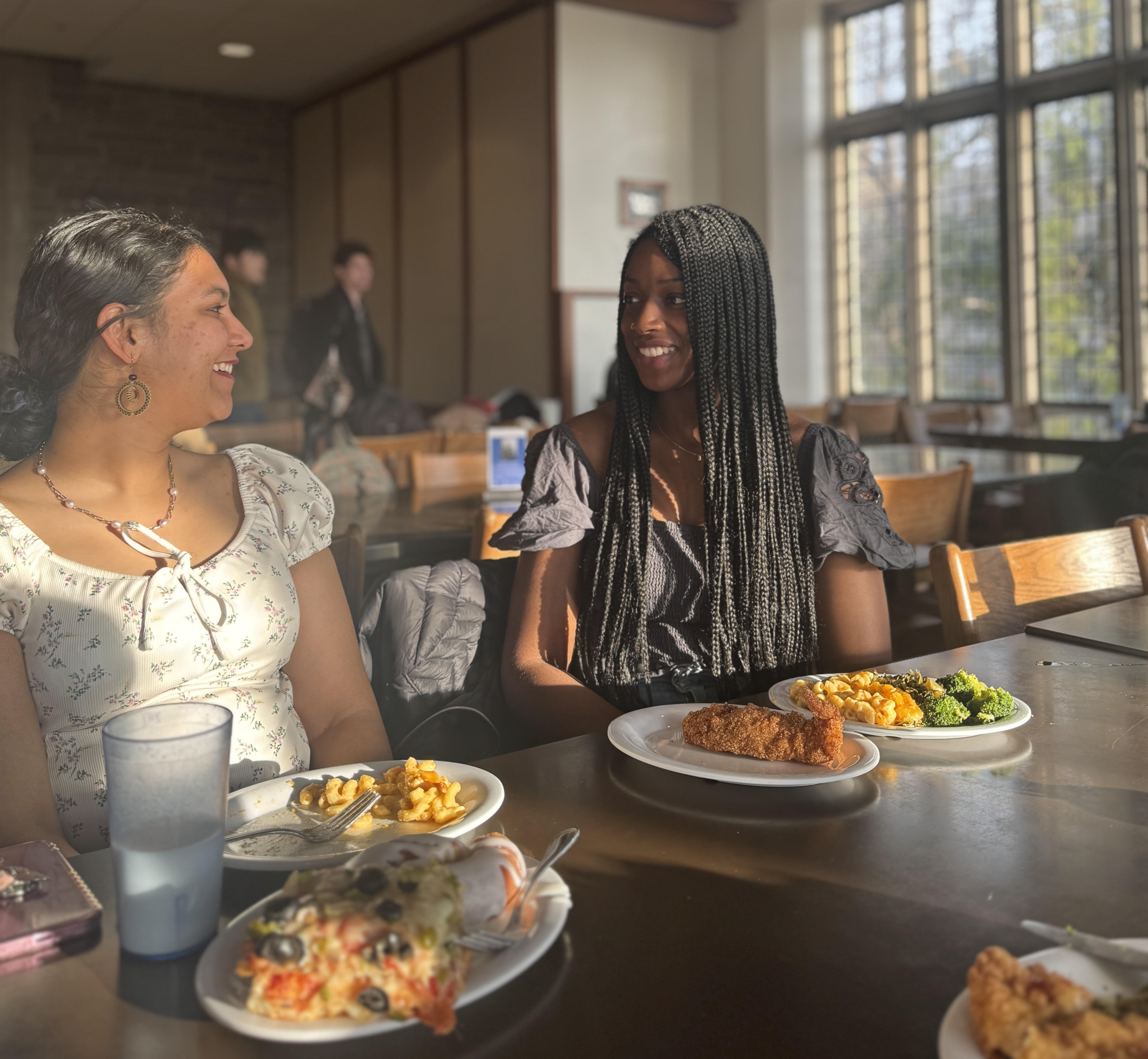 Two students smile at each other at a table with plates of food in front of them