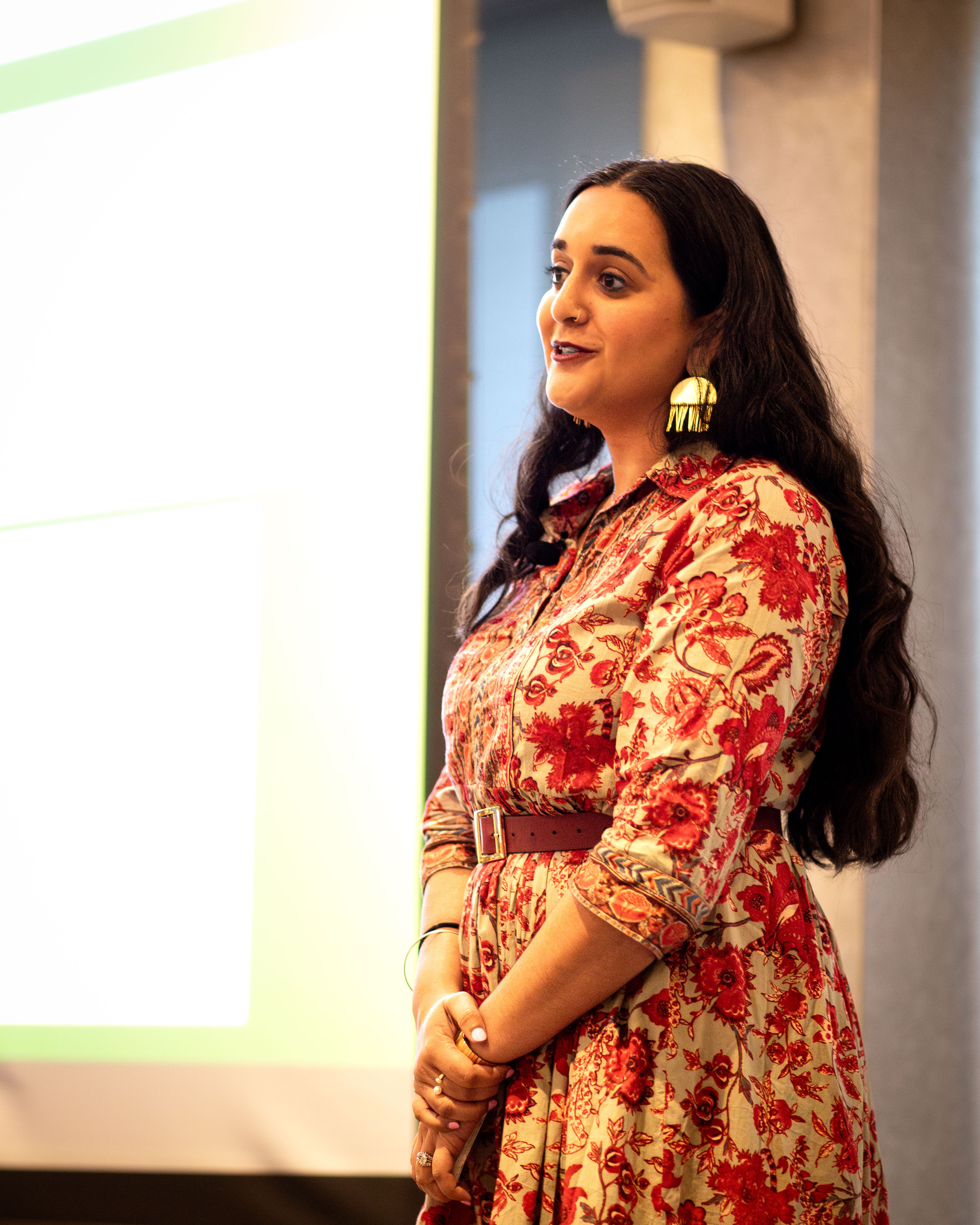 A woman in a floral red and beige dress speaks in front of a projected screen at the APIDA Gala, wearing gold statement earrings and standing with her hands clasped.