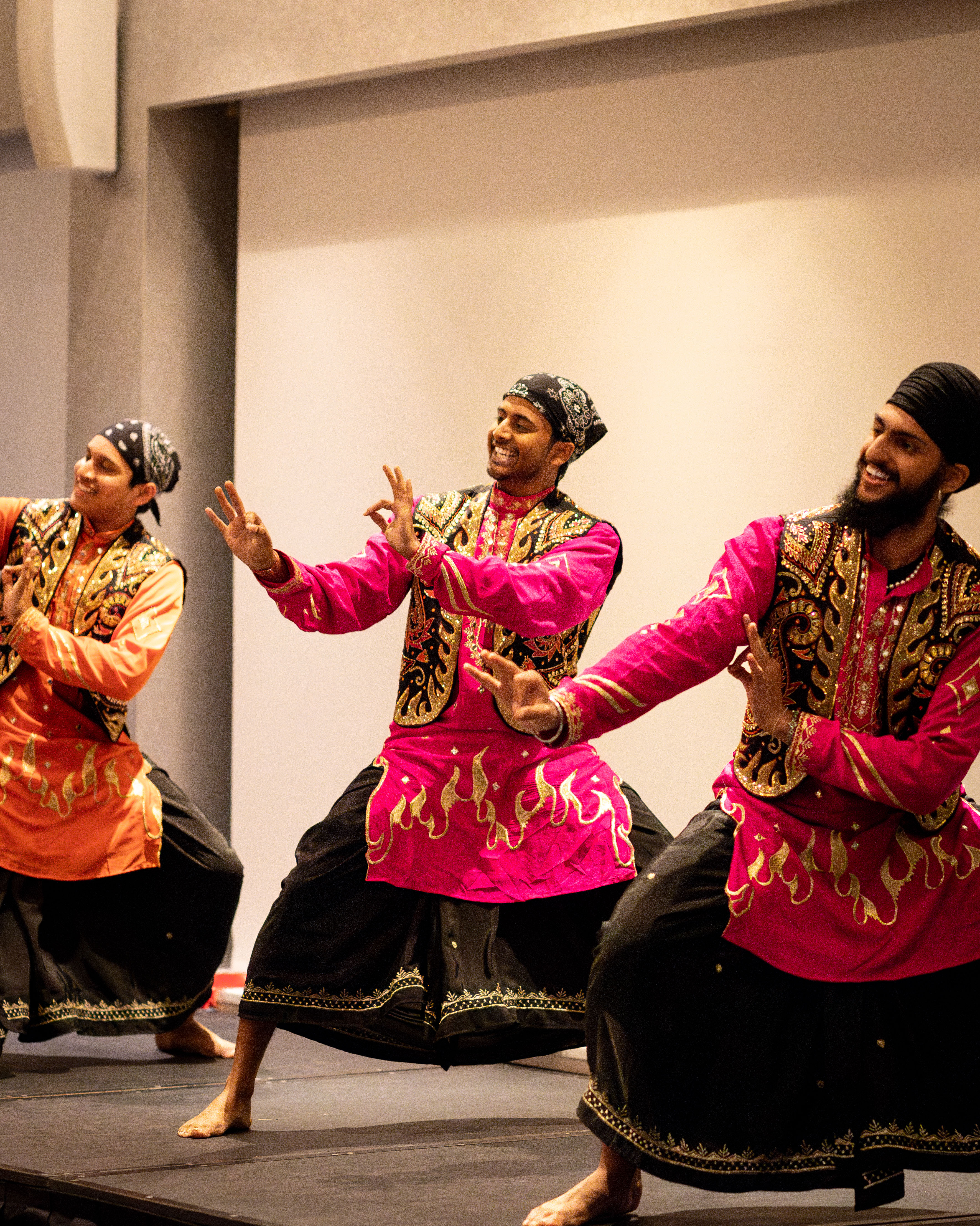 Three male dancers perform a traditional South Asian dance on stage, wearing vibrant costumes with black pants and embellished pink and orange tops, expressing joyful energy through dynamic hand and body movements.
