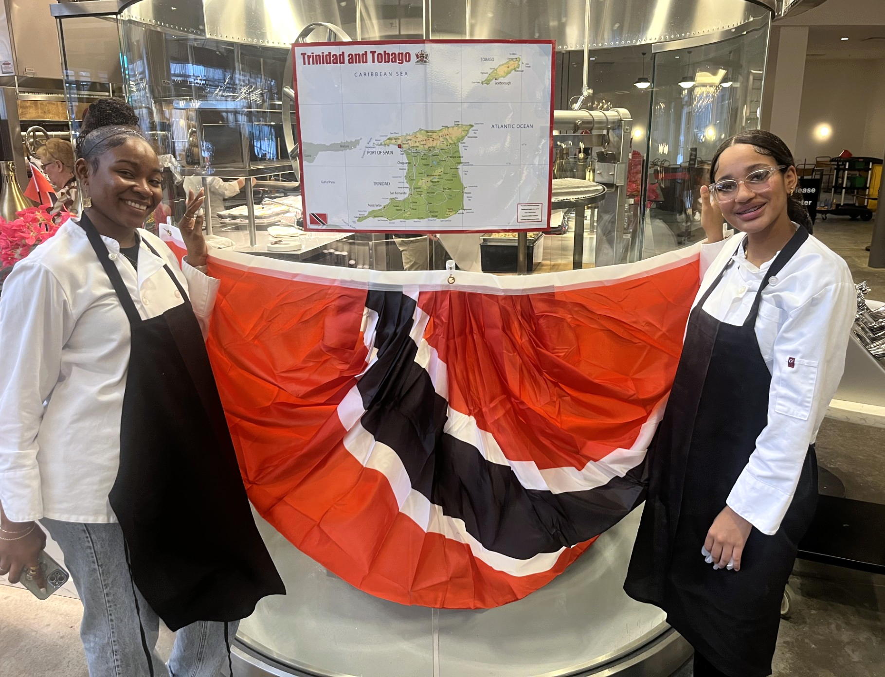 Two students in aprons hold up a Trinidadian flag