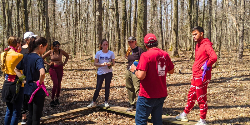 Group of students standing at a low element while a challenge course facilitator in a red shirt leads a discussion.