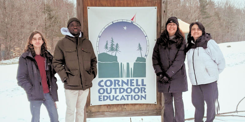 Four student coordinators standing outside in the snow next to a Cornell Outdoor Education sign