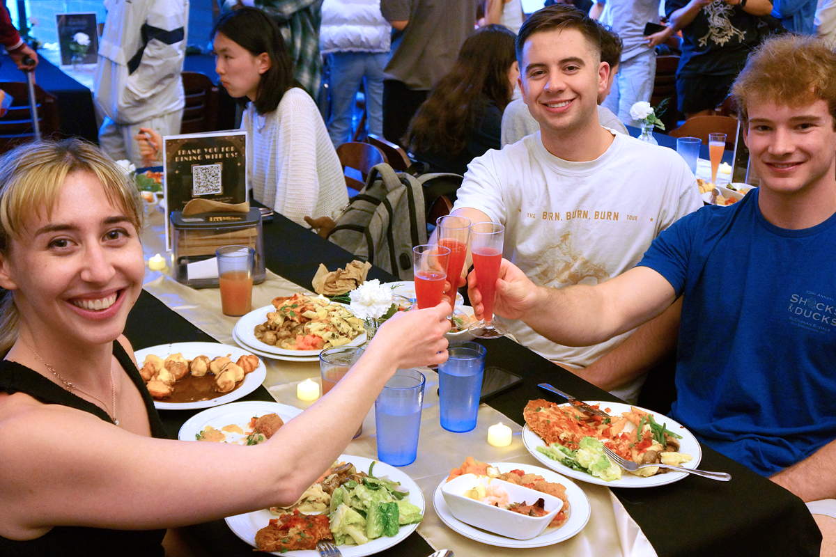 Three people at a table with plates of food clink glasses together in a toast