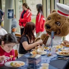 Students sit at a dining room table with plates of food. One of them is chatting with a bear mascot wearing a chef outfit