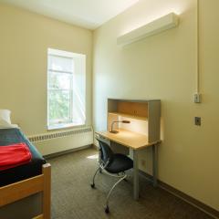 Single room with a twin bed, desk, chair, lamp, dresser, and window.