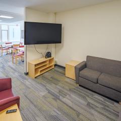A large lounge in Cascadilla Hall with wall mounted tv, couches, tables and chairs.
