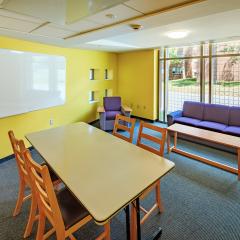 A lounge in Loving House with a long table, four chairs, a couch and lounge chair facing a coffee table, a white board on the yellow wall and a large window. 