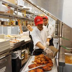 A person in a kitchen takes fried chicken out of a fryer basket, with two others looking on