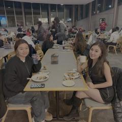 Two young women sit at a dining room table holding drinks, with plates of food in front of them