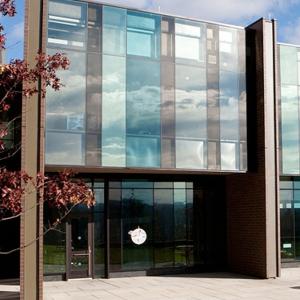 The facade of a college residential building with vertical girders and glass walls. A tree is in front, and there are picnic tables outside