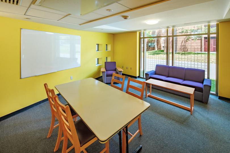 A lounge in Mews Hall with a long table, four chairs, a couch and lounge chair facing a coffee table, a white board on the yellow wall and a large window. 