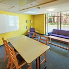A lounge in Mews Hall with a long table, four chairs, a couch and lounge chair facing a coffee table, a white board on the yellow wall and a large window. 