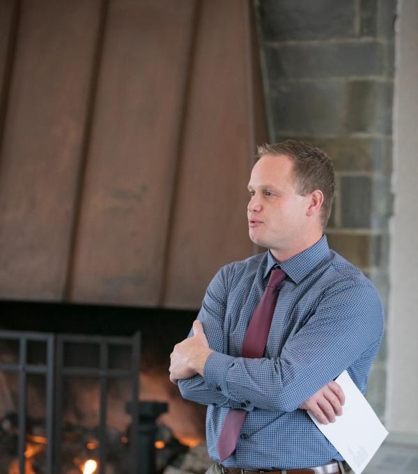 A man wearing a tie speaking with arms folded. A fireplace is in the background.