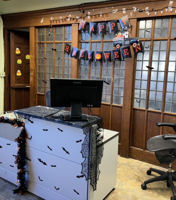 A desk and rolling office chair in front of a wood and frosted glass wall. Halloween decorations include a Happy Halloween banner on the wall, and a spiderweb drape and paper bats on the desk