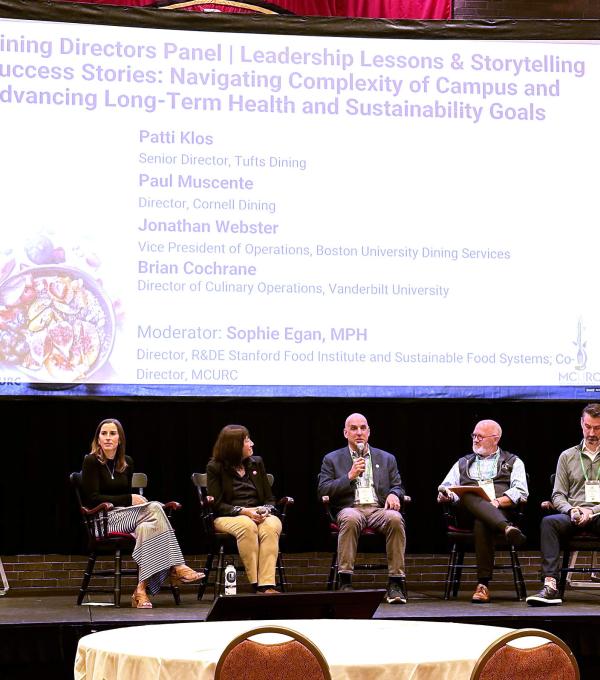 Five people sit in chairs on a stage in front of a projector screen, with one speaking into a microphone