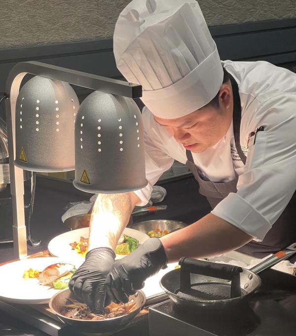 A person wearing a chef jacket and hat and apron bends over a cooking station plating the food