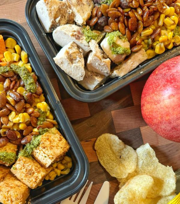 Two takeout containers full of food, with potato chips and an apple on the table next to them