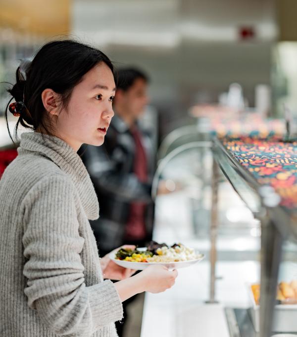 A young woman stands in front of a food serving station with a plate