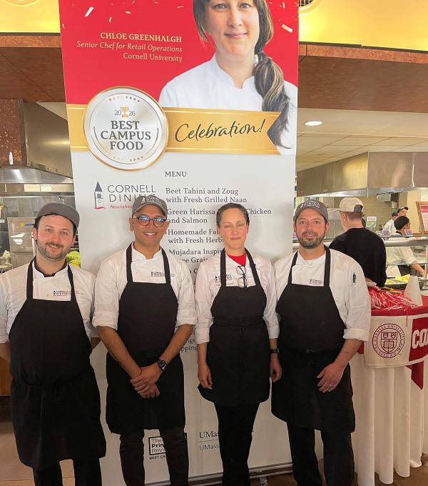 Four people in chef coats and aprons in front of a poster with a woman's face